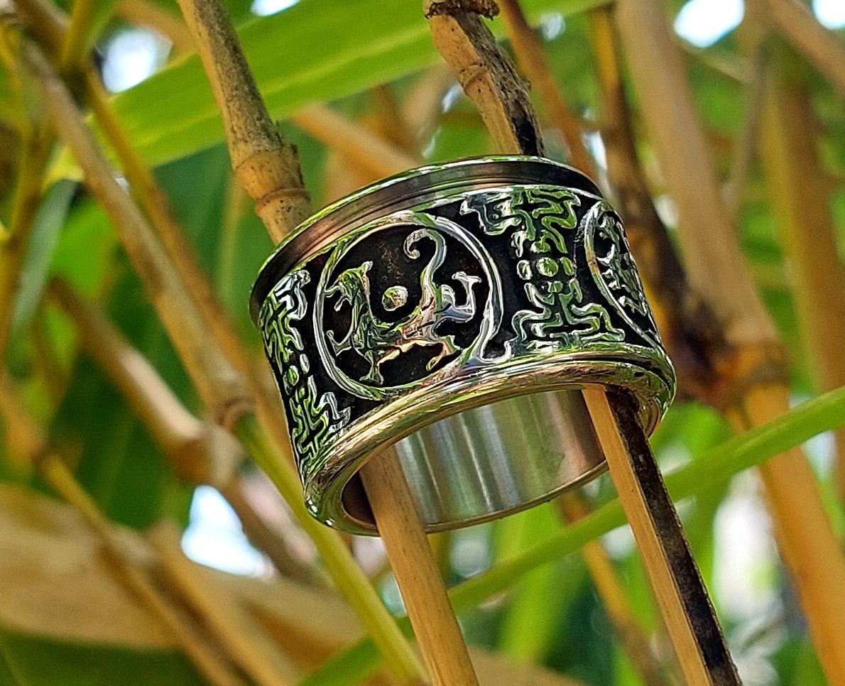 A wide silver-toned metal ring with black enamel and raised green dragon motifs, shown hanging from bamboo branches in a natural outdoor setting.