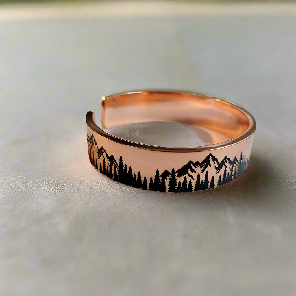 Rose gold cuff bracelet with mountain and tree design on a white surface with keyboard in the background