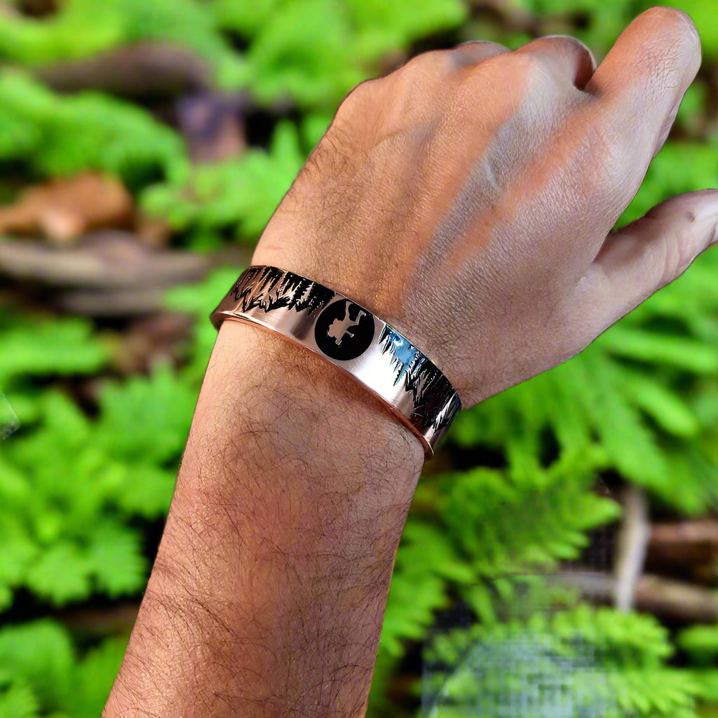 Hand wearing a metallic bracelet with a forest design, sitting on a desk.