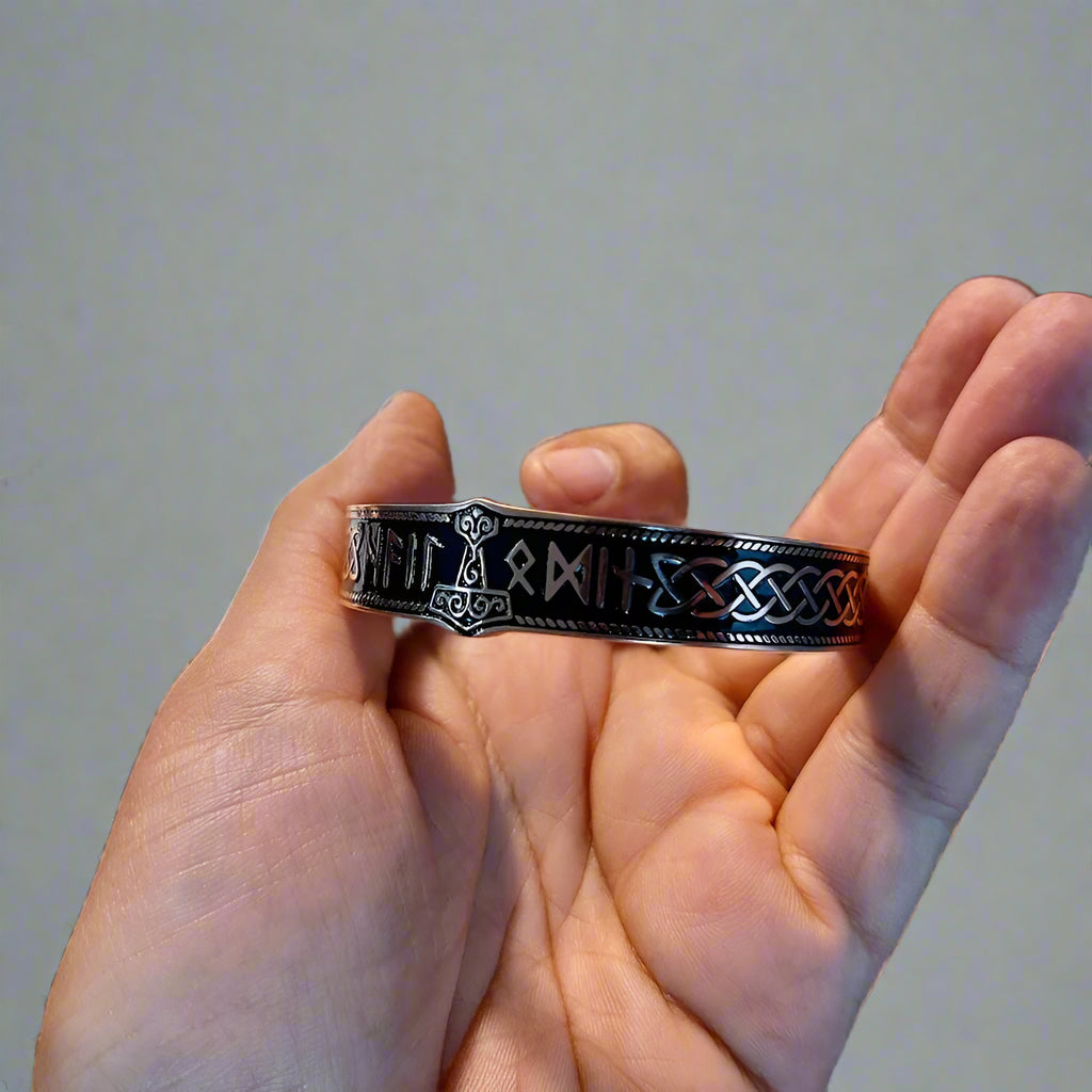 Hand holding a metallic bracelet with intricate designs on a dark surface.