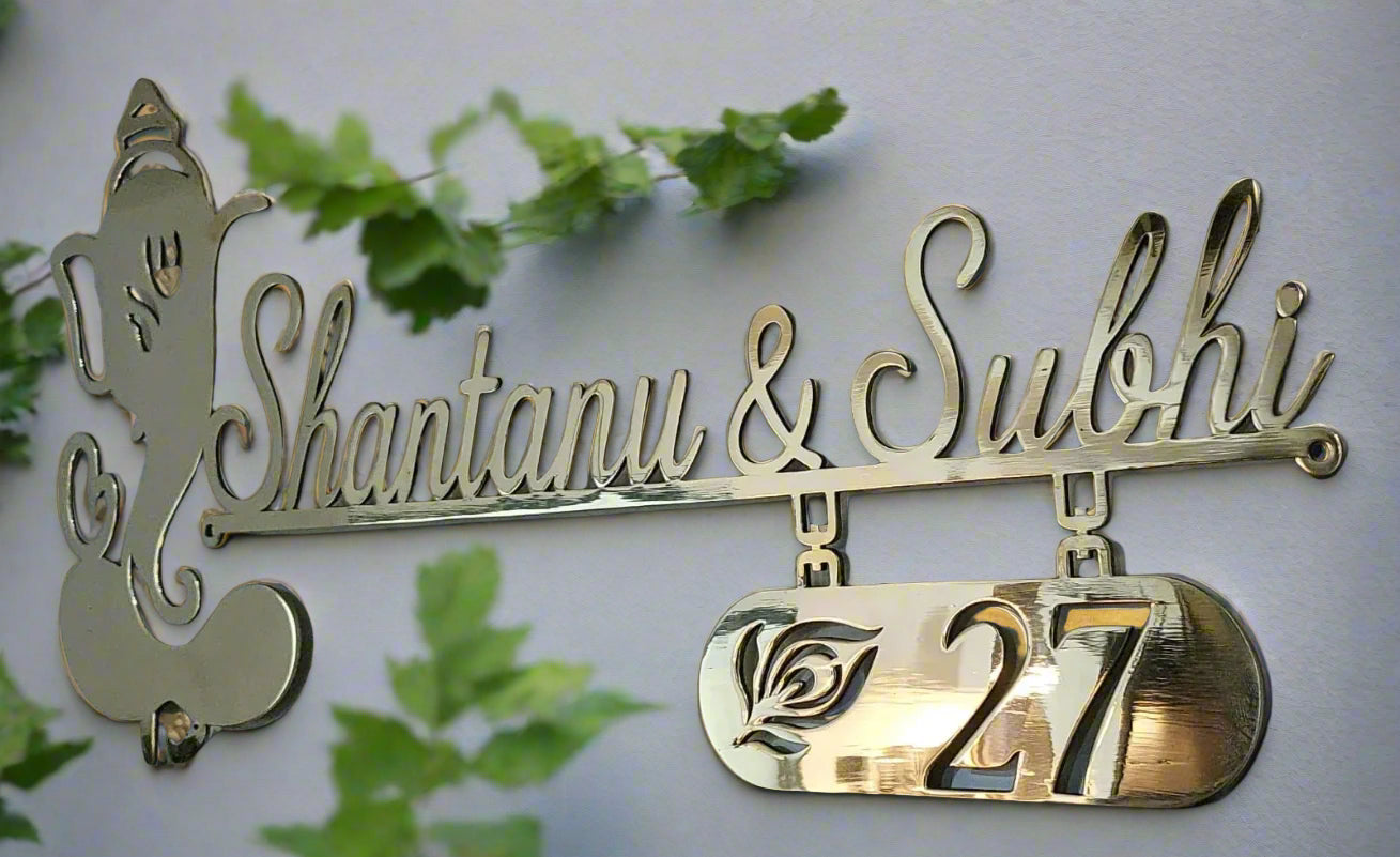Decorative nameplate with 'Shantanu & Subhi' and number '27' on a light background with green leaves.